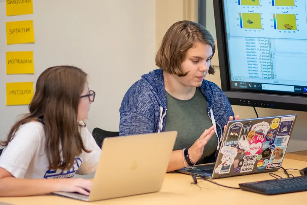 Two students sit at a table next to each other while working on lap tops.